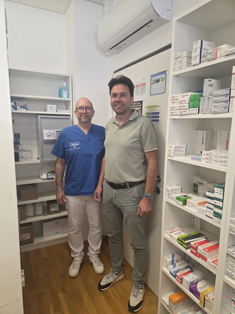Dr. Lechner und ein Mann stehen in einer Apotheke mit Regalen voller Medikamente.