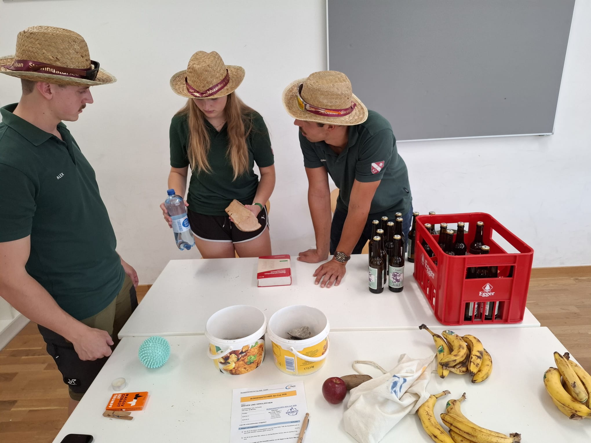 Drei Personen mit Strohhüten und grünen Poloshirts stehen an einem Tisch mit Bierflaschen, Bananen und Behältern.