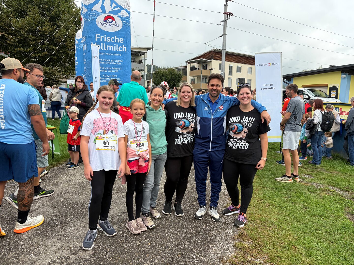 Läufer und Kinder mit Medaillen bei einem Lauf in Österreich, im Hintergrund eine Milchwerbung.
